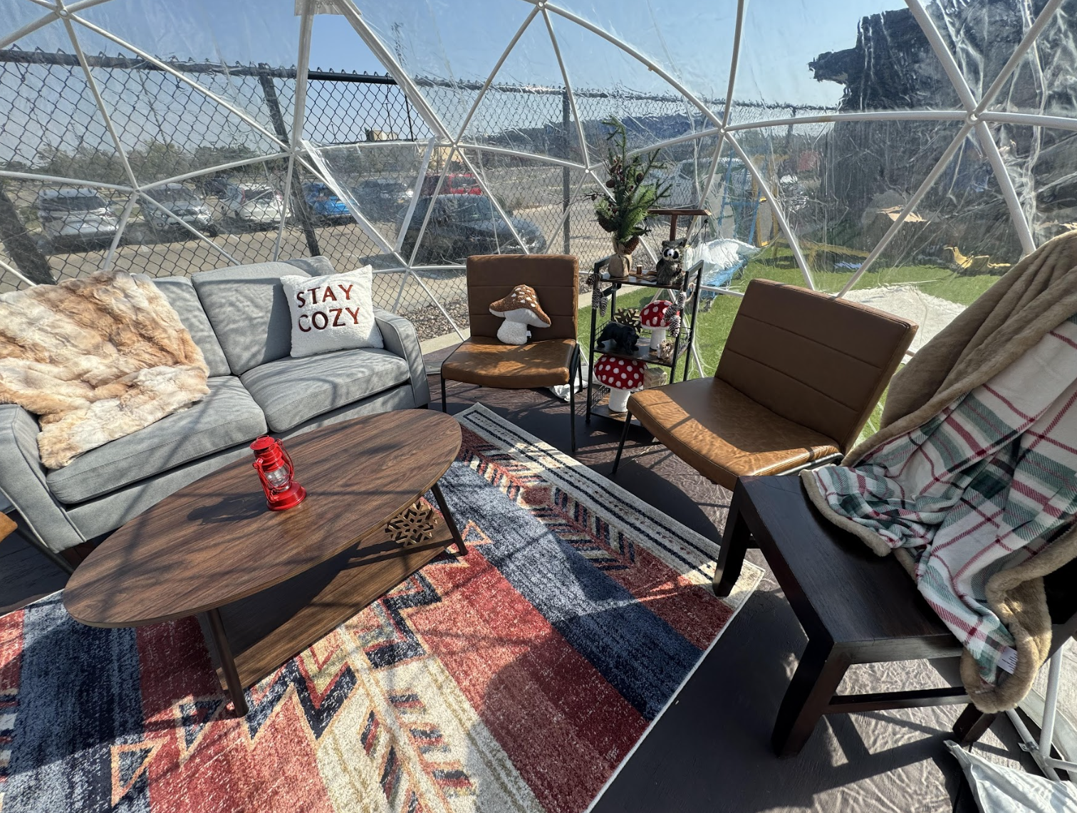 A cozy dome tent with a gray couch, two brown chairs, decorative pillows, a throw blanket, a wooden coffee table, and a patterned rug. Sunlight streams in, and a chain-link fence is visible outside.